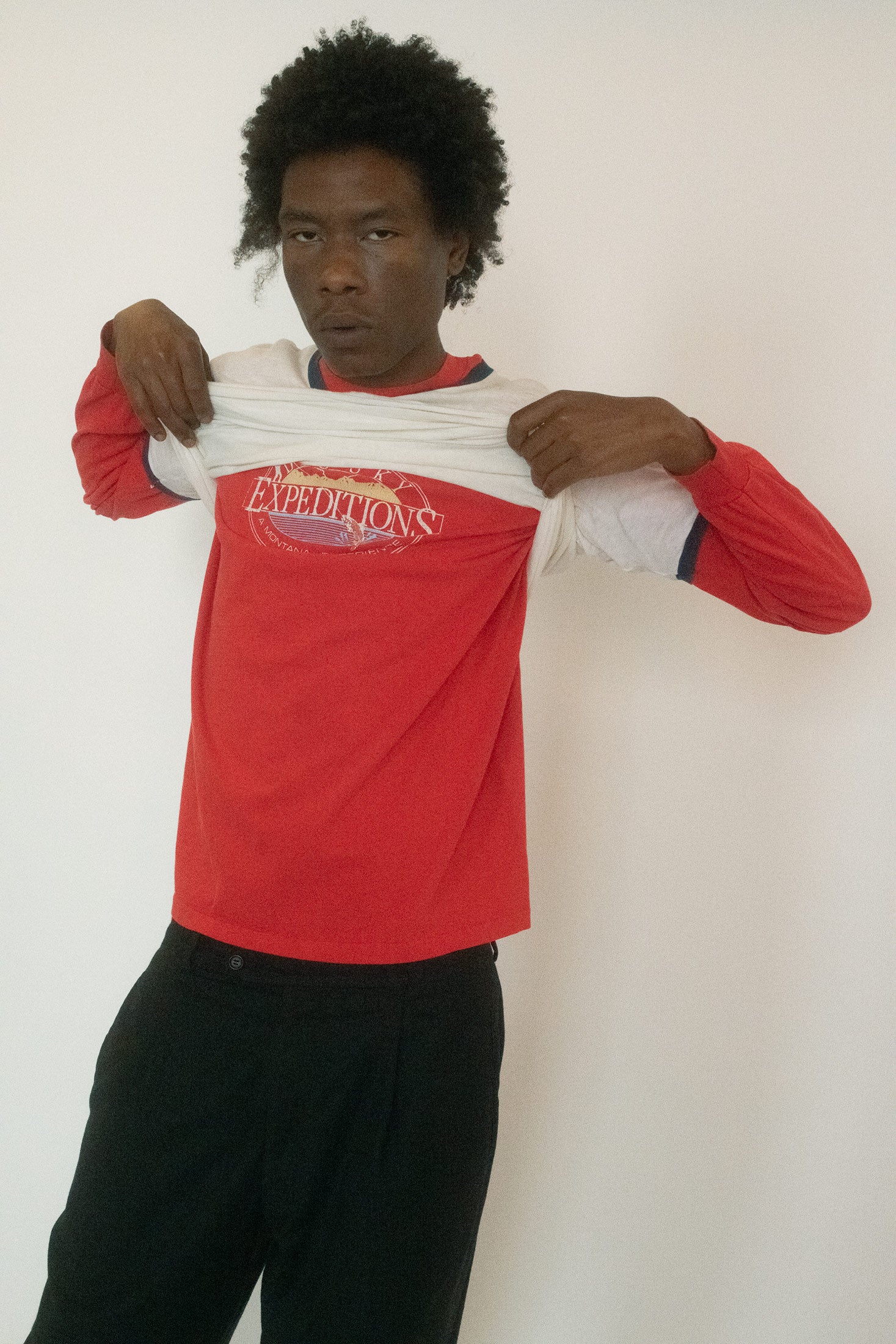 The ringer tee, casual and cropped in attitude, plays against the saturated red sleeves beneath, the colors clashing just enough to feel deliberate.