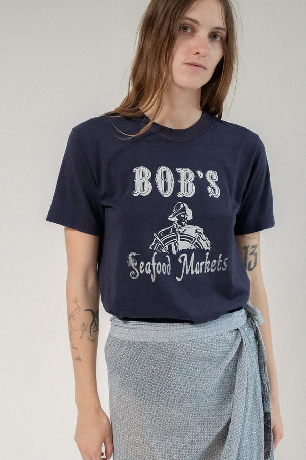 The night was thick with neon and static. The Vintage Bob’s Tee moved through it like it belonged there—navy, faded just right, with white text that caught the streetlight like a smirk.