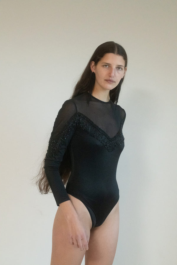 The city is alive with whispers, and you, poised in the shadows, are the secret they’ll never forget. The Giorgio Sant'Angelo beaded bodysuit hugs your form like a second skin, the black shiny fabric reflecting the neon lights as you move through the night. 